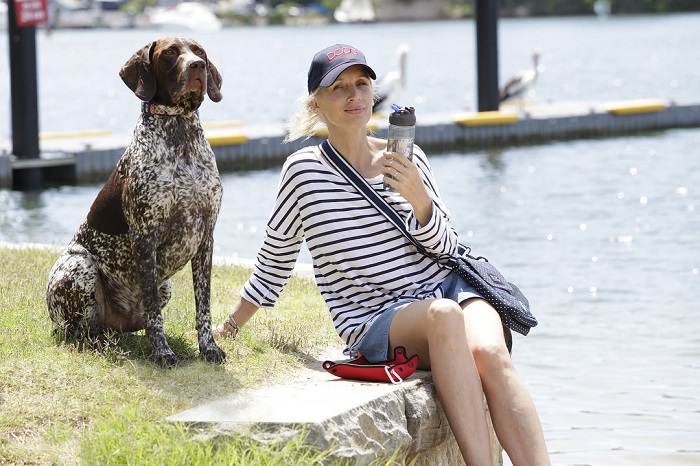 Woman wearing DOOG brand dog walking accessories and hat sitting with a German Shorthaired Pointer by a lake.