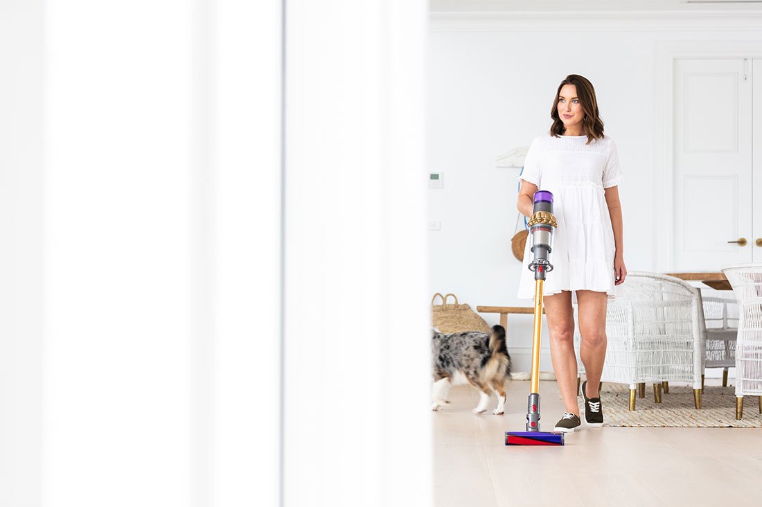 Woman using a Dyson V11 vacuum to clean pet hair from hardwood floors in a home with a dog.