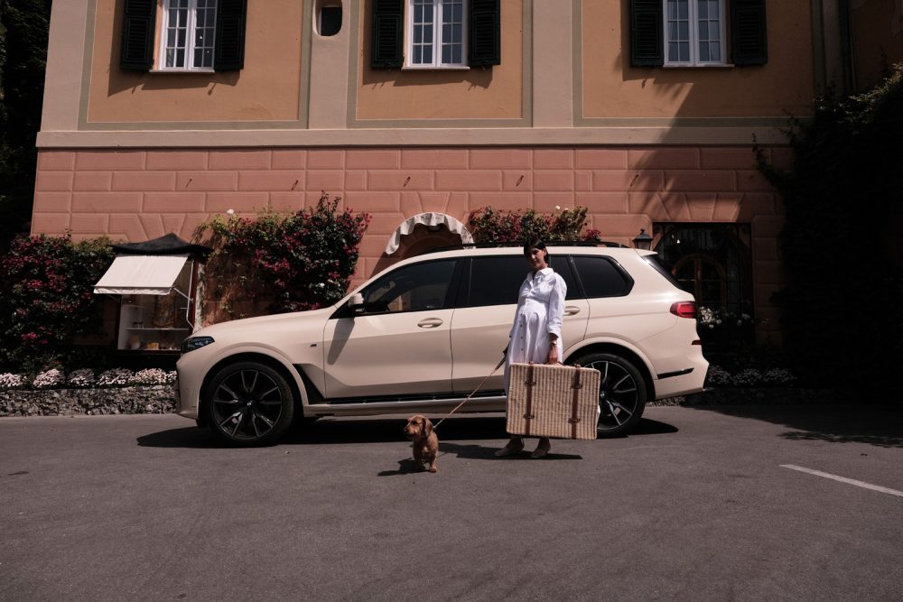 Woman with dachshund and wicker trunk by a BMW SUV for the BMW Italia x Poldo Dog Couture luxury pet travel collection.