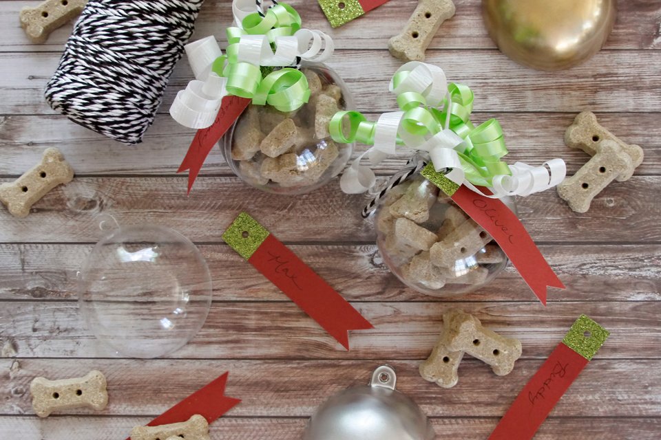 DIY dog treat bauble ornaments filled with bone-shaped biscuits, featuring green ribbons and personalized name tags.