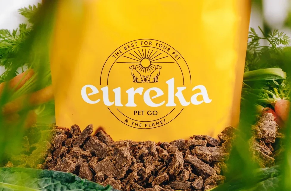 Eureka Pet Co yellow bag of Australian air dried dog food surrounded by fresh vegetables like carrots and kale.