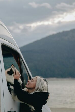 A dog mum petting her husky looking out of a camper van window while RV camping with her dog by a scenic lake.