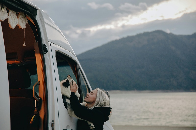 A dog mum petting her husky looking out of a camper van window while RV camping with her dog by a scenic lake.